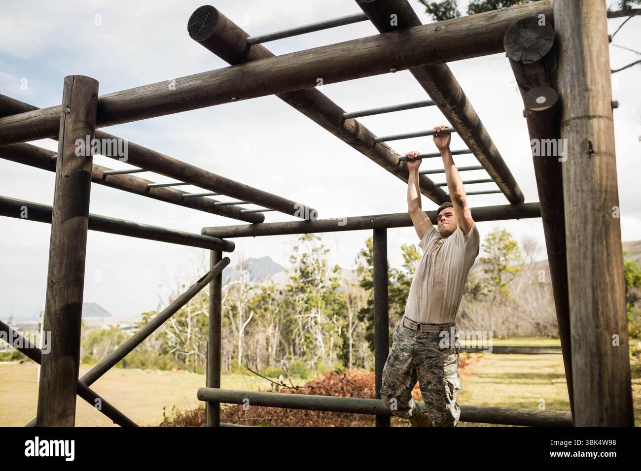 Soldato maschio con cintura di sicurezza che naviga sulle barre delle scimmie su un terreno di allenamento erboso sotto il cielo luminoso Foto Stock