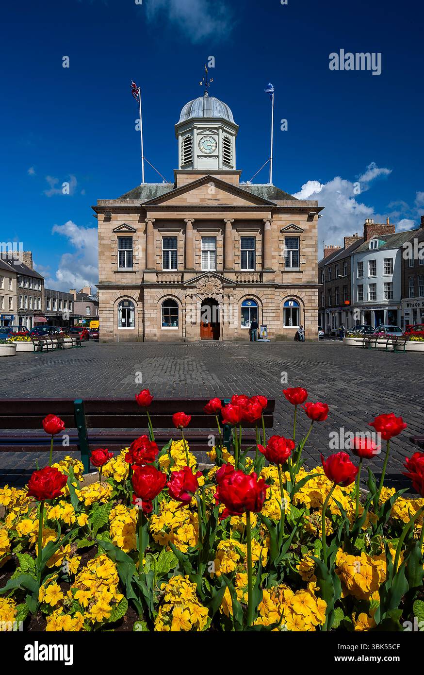 Una vista in una giornata di sole d'estate del municipio di Kelso Town Square, nei confini scozzesi, con gente che macina intorno e mostre di fiori Foto Stock
