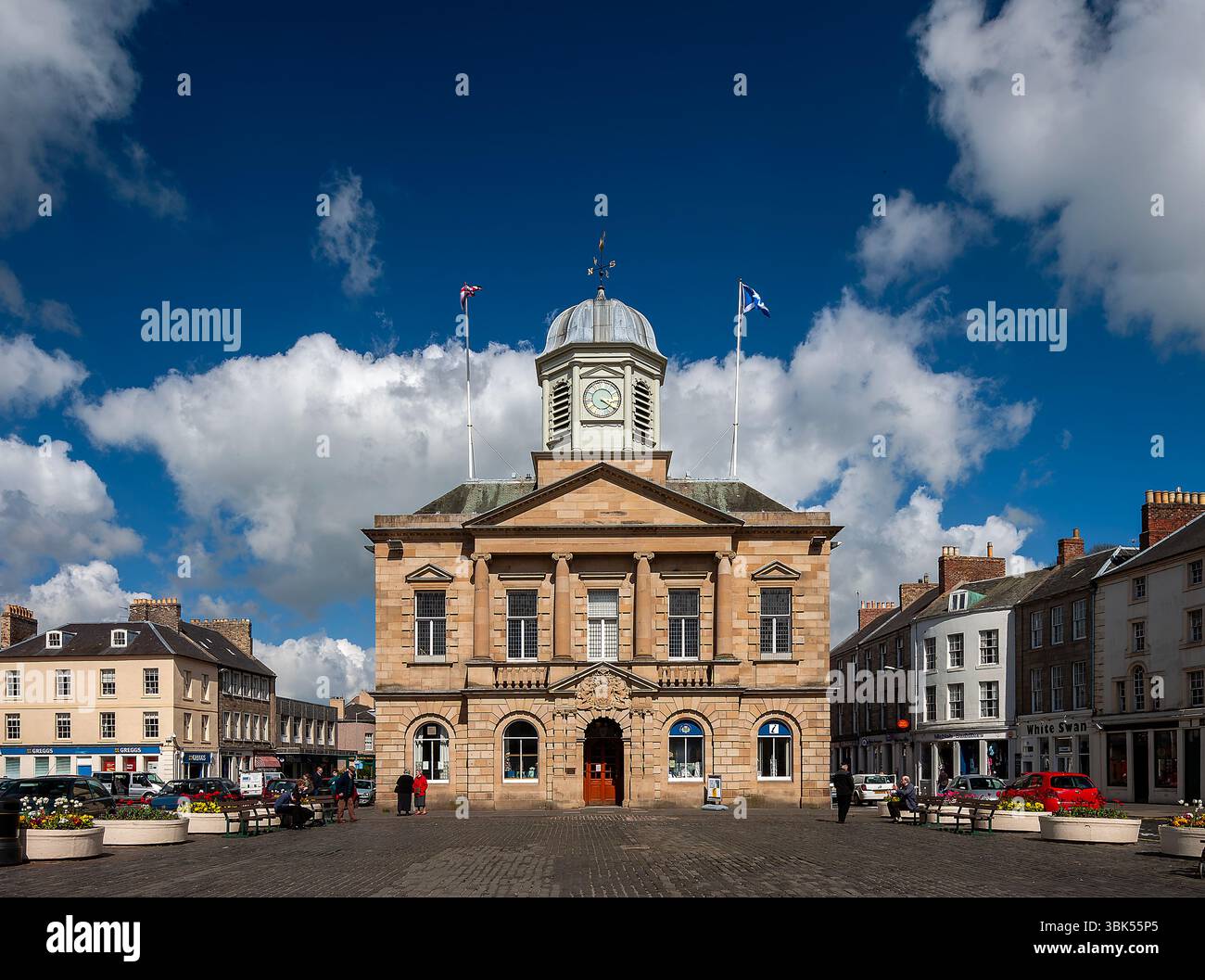 Una vista in una giornata di sole d'estate del municipio di Kelso Town Square, nei confini scozzesi, con gente che macina intorno e mostre di fiori Foto Stock