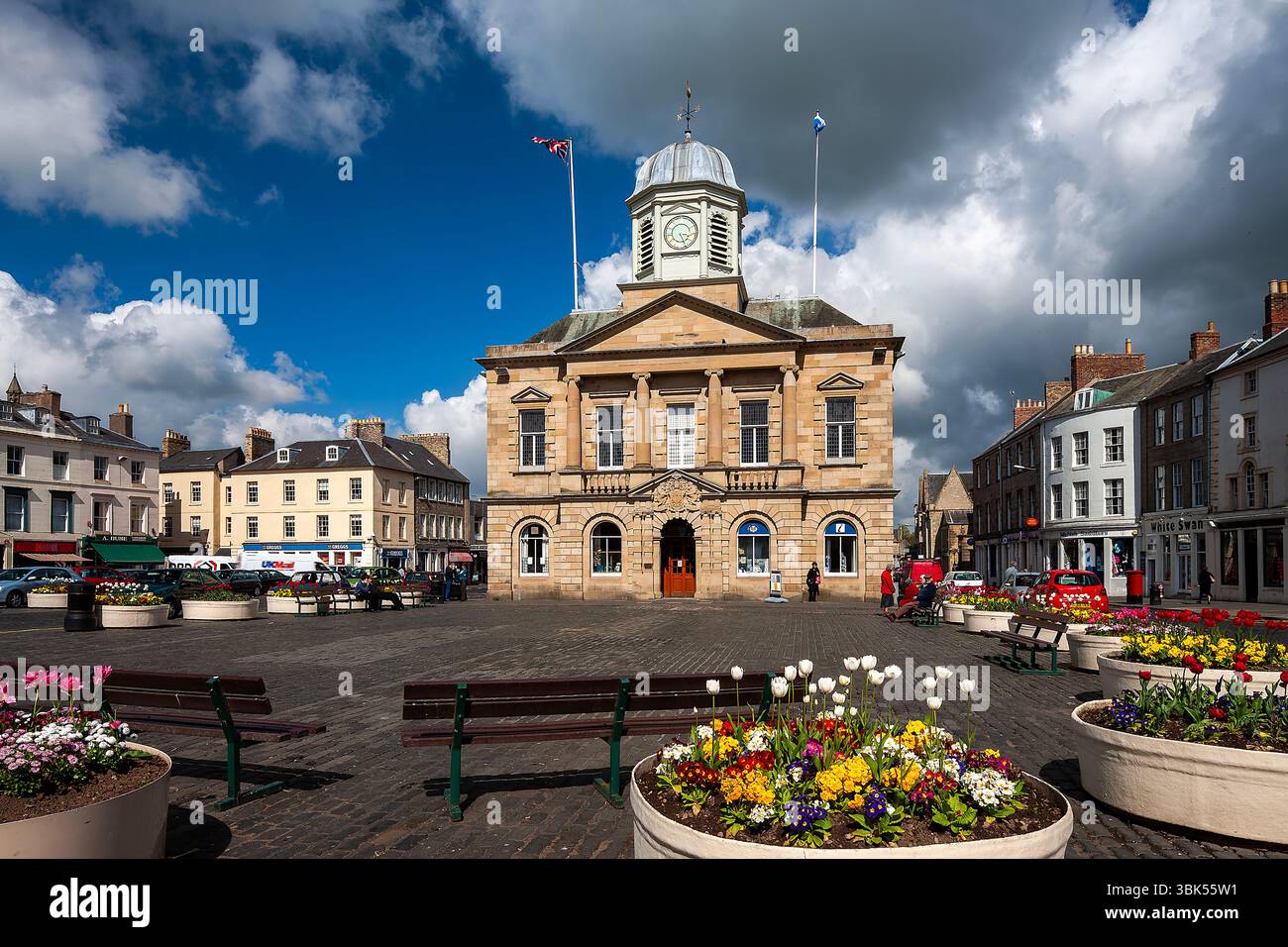 Una vista in una giornata di sole d'estate del municipio di Kelso Town Square, nei confini scozzesi, con gente che macina intorno e mostre di fiori Foto Stock