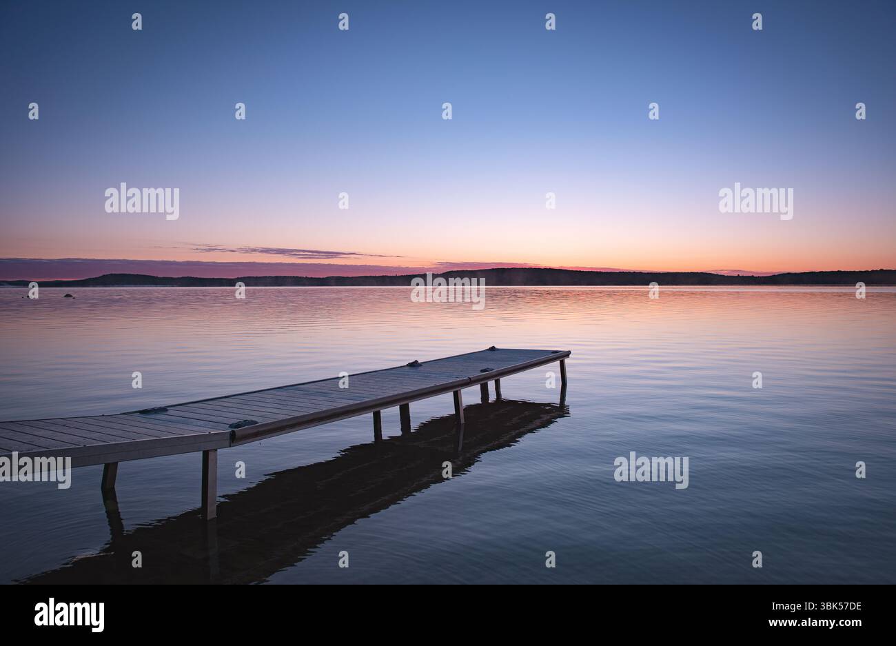 Un molo si estende in un lago ancora settentrionale all'alba, riflettendo i colori tenui dell'alba. Ambiente naturale tranquillo e remoto alla luce del mattino presto. Foto Stock