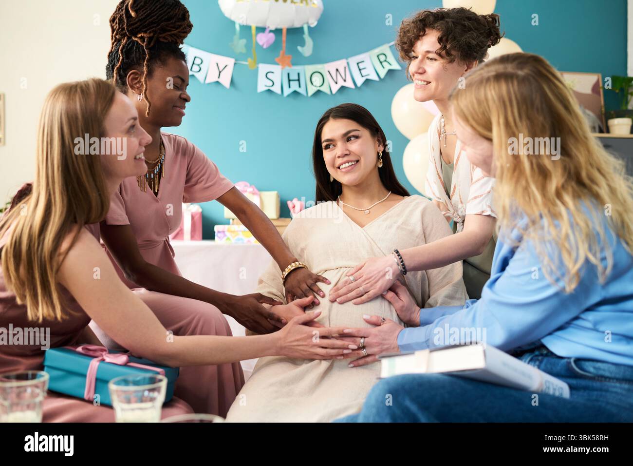 Gruppo multietnico di giovani donne adulte che circondano una giovane donna adulta incinta, sorridendo e toccando la pancia durante la festa di rivelazione di genere Foto Stock