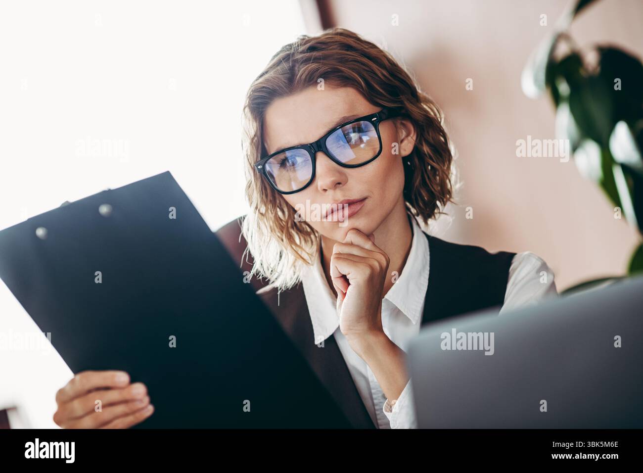 Donna d'affari sicura che rivede i documenti in uno spazio di lavoro professionale con un'espressione contemplativa e un abbigliamento formale Foto Stock