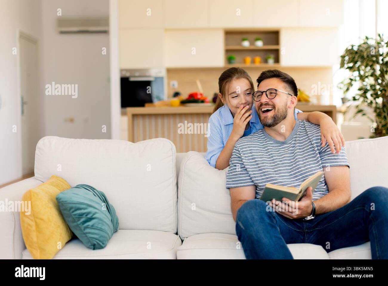 Un padre sorridente condivide una storia deliziosa con sua figlia, seduto su un comodo divano in una casa luminosa e moderna. Foto Stock