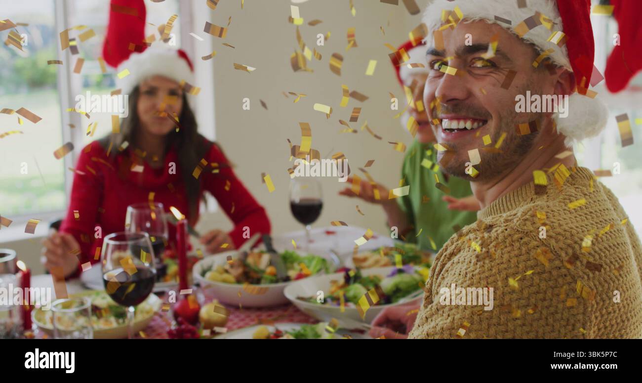 Riunire gli amici che indossano i cappelli di Babbo Natale intorno al tavolo da pranzo di casa, con bicchieri da vino e coriandoli d'oro Foto Stock