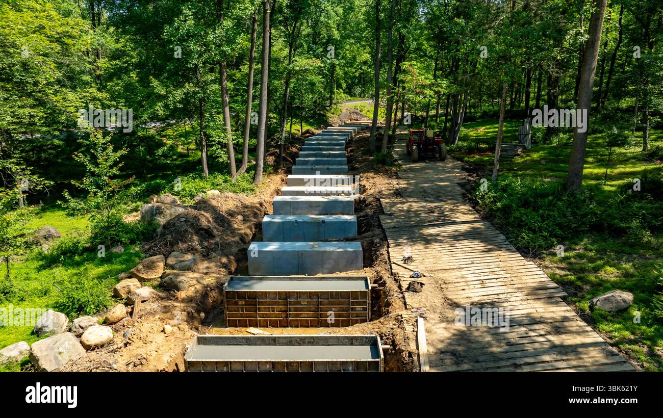 È in corso la costruzione di un nuovo sentiero in un'area boschiva verde. Le tavole di legno fanno parte del percorso, mentre le forme di calcestruzzo sono disposte in modo organizzato. Sullo sfondo sono presenti macchinari pesanti. Foto Stock