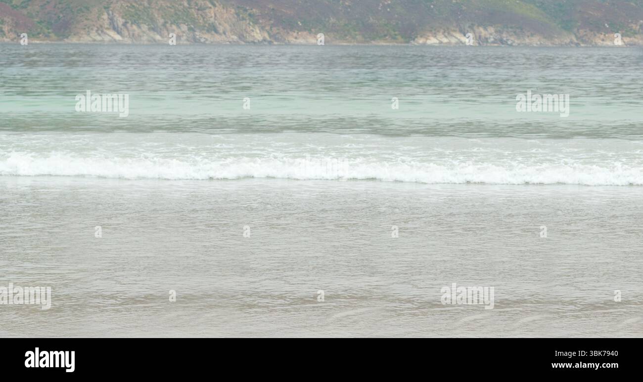 Le onde d'acqua poco profonde si stagliano sulla spiaggia sabbiosa in un design piatto, cielo coperto, colline rocciose Foto Stock