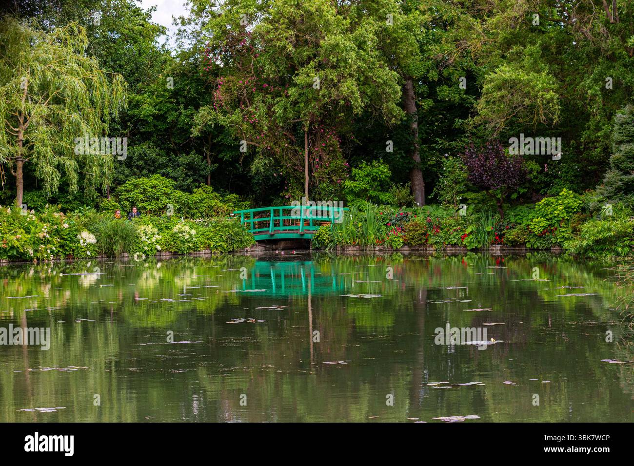 Il verde rigoglioso incornicia un laghetto riflettente con una passerella verde ad arco sullo sfondo a Giverny, Francia. Foto Stock