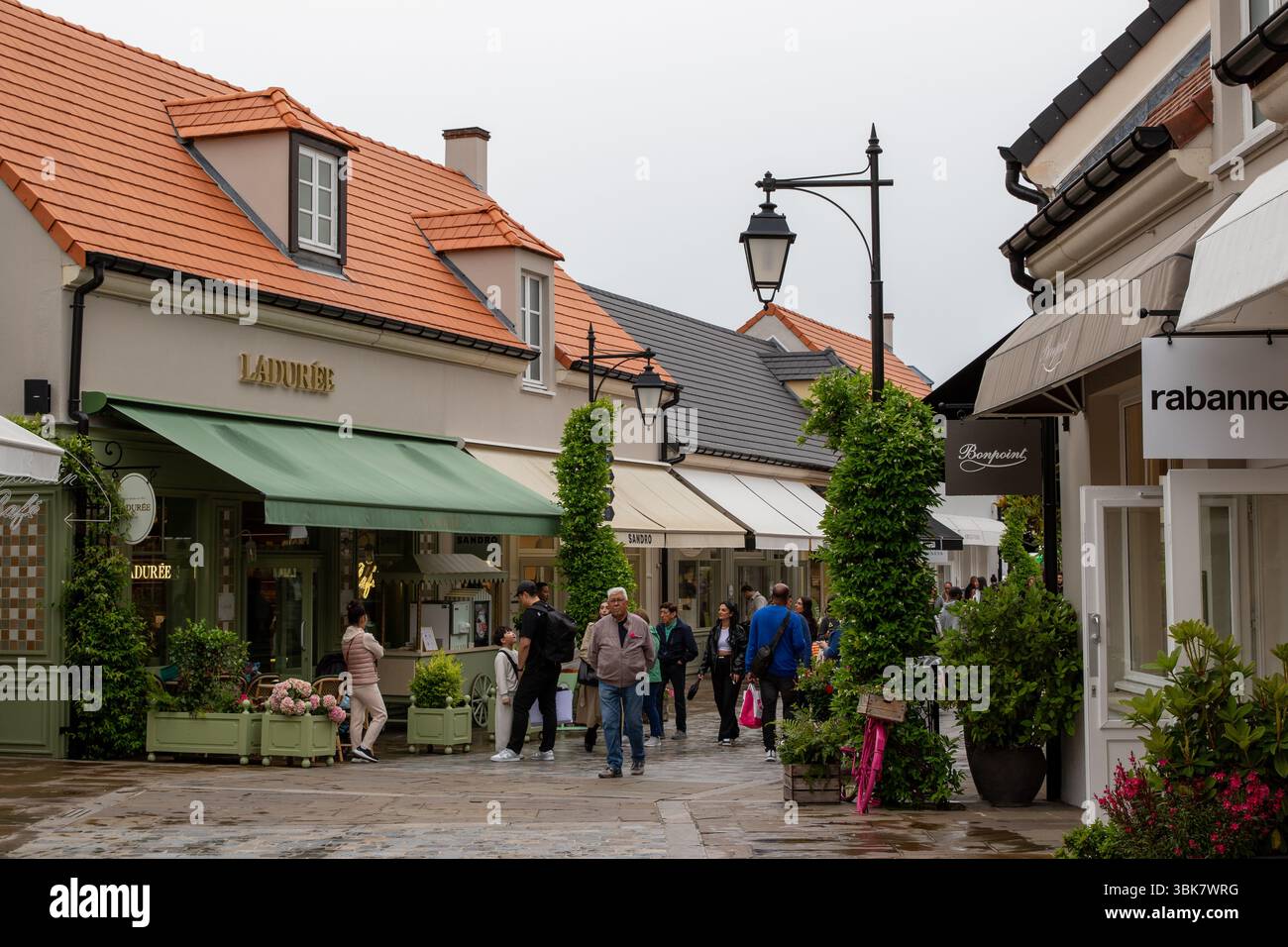 Persone che camminano su una strada fiancheggiata da vetrine, sotto un cielo nuvoloso. Gli edifici hanno tetti arancioni e pareti beige al la Vallee Village, in Francia. Foto Stock
