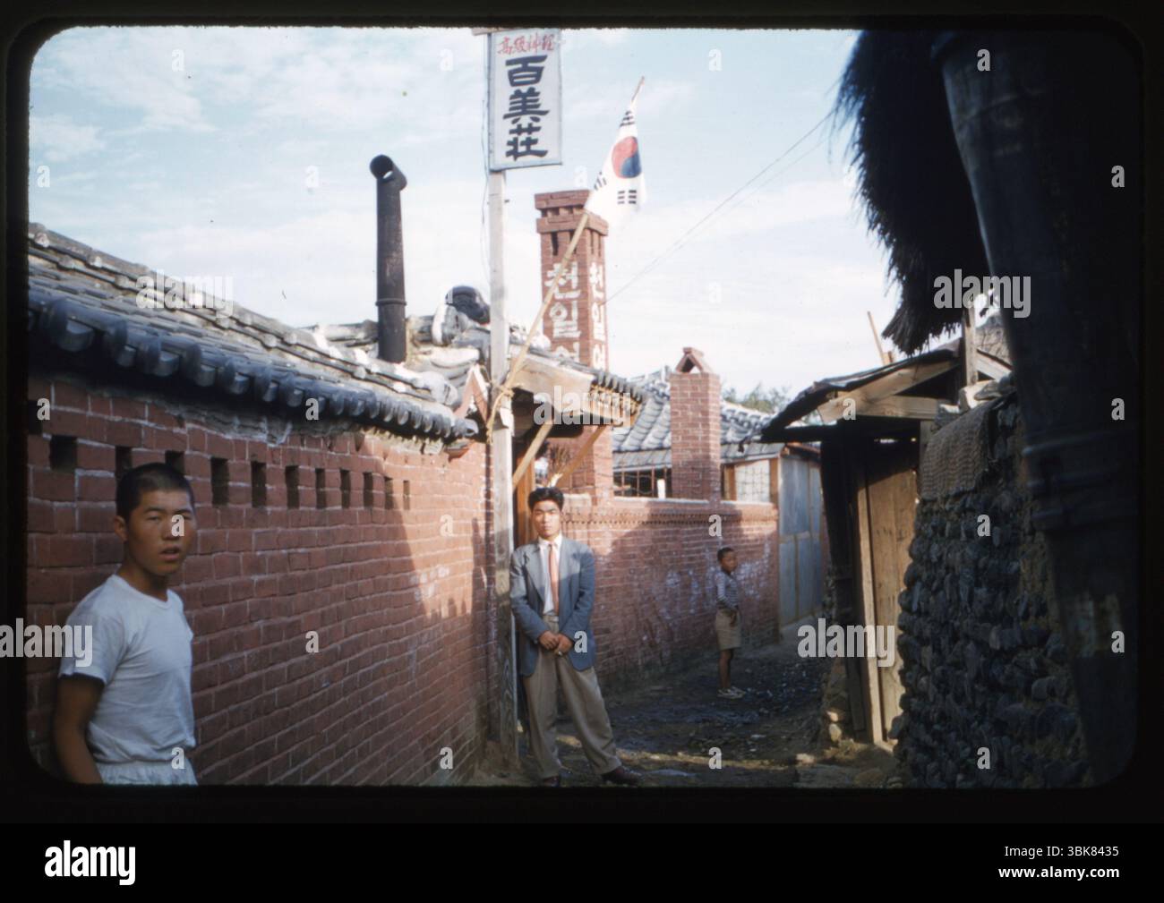 Una vista sulla strada fuori da una casa Kisang (geisha coreana) a Masan, Corea, ottobre 1956. Uomini e ragazzi si trovano in un vicolo fiancheggiato da case in mattoni e pietra con tradizionali tetti piastrellati. Sono visibili la bandiera nazionale coreana e numerosi segni a Hangul, compresa la parola "천일여관" (locanda di Cheonil). Catturata da un militare statunitense durante l'occupazione post-bellica. Questa fotografia è stata scattata dal Chief Petty Officer Wilfred K.C. Park, un coreano americano che prestava servizio nella Marina degli Stati Uniti, che ha documentato la vita e le operazioni militari in Corea e Giappone durante gli anni '1950 Foto Stock