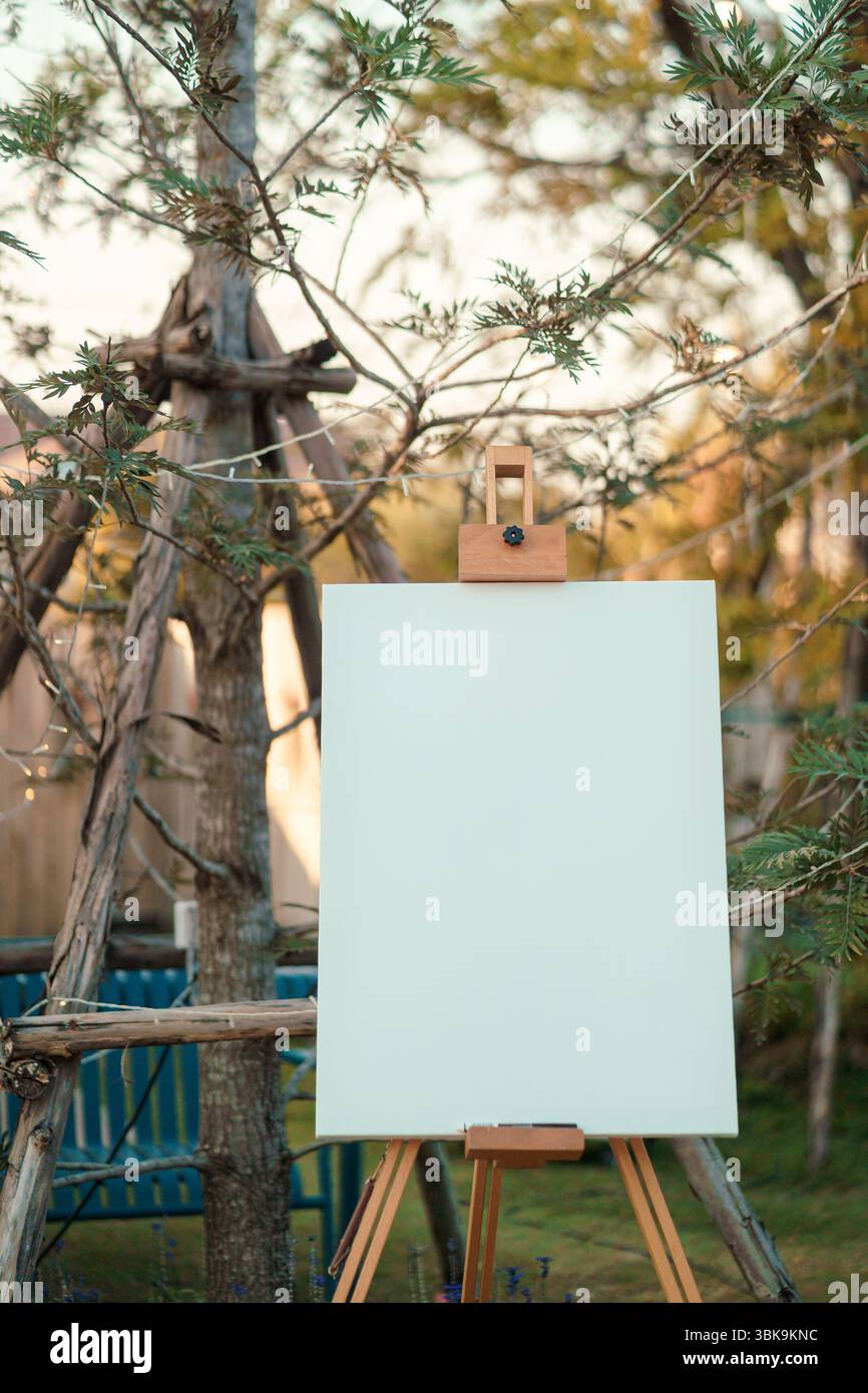 Tela bianca bianca su cavalletto in legno collocata di fronte all'evento nuziale in un ambiente naturale e in un morbido giardino in stile rustico Foto Stock
