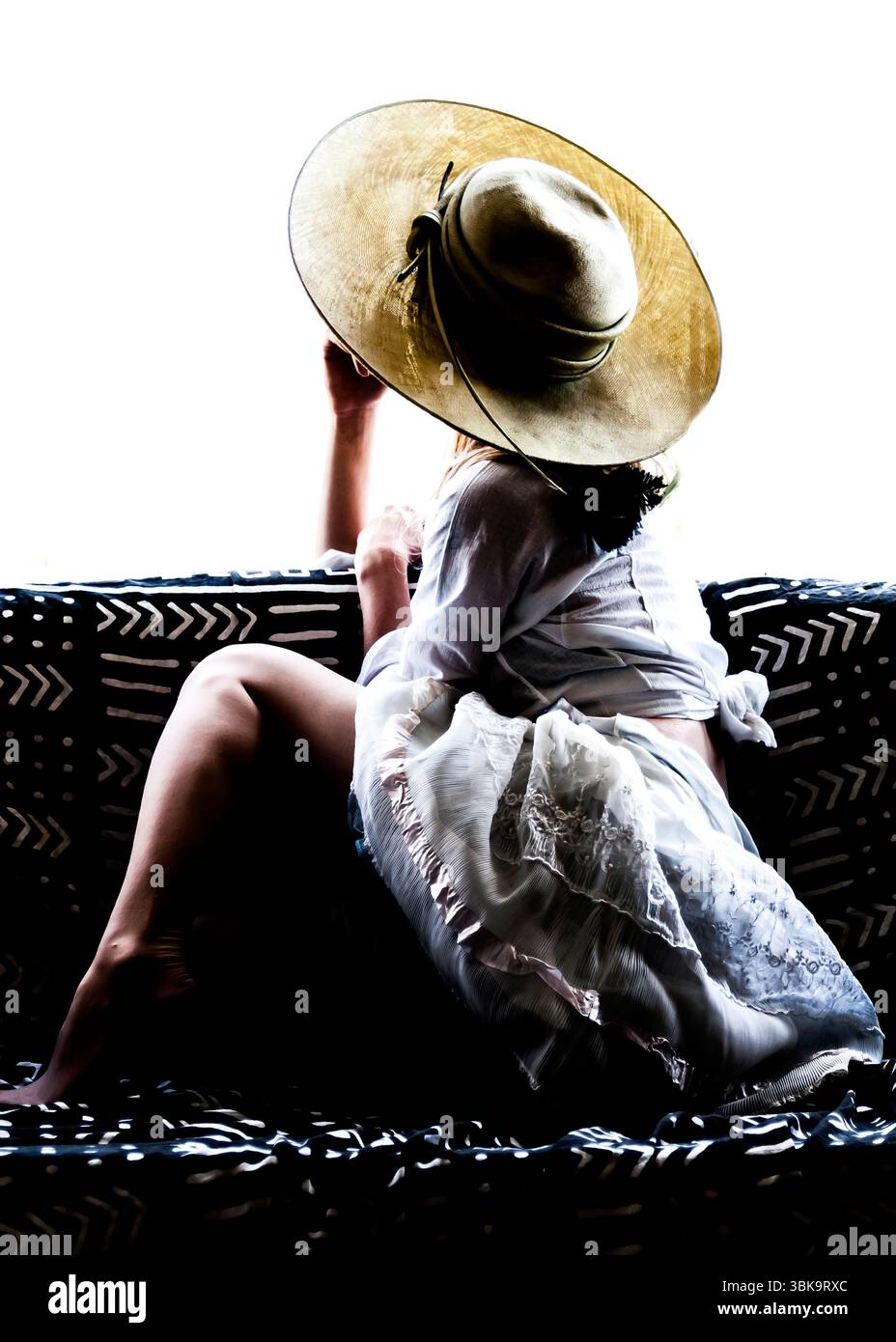 Una donna che indossa un grande cappello si siede su un divano a motivi geometrici, il suo volto oscurato. Sta indossando un vestito di colore chiaro. Foto Stock