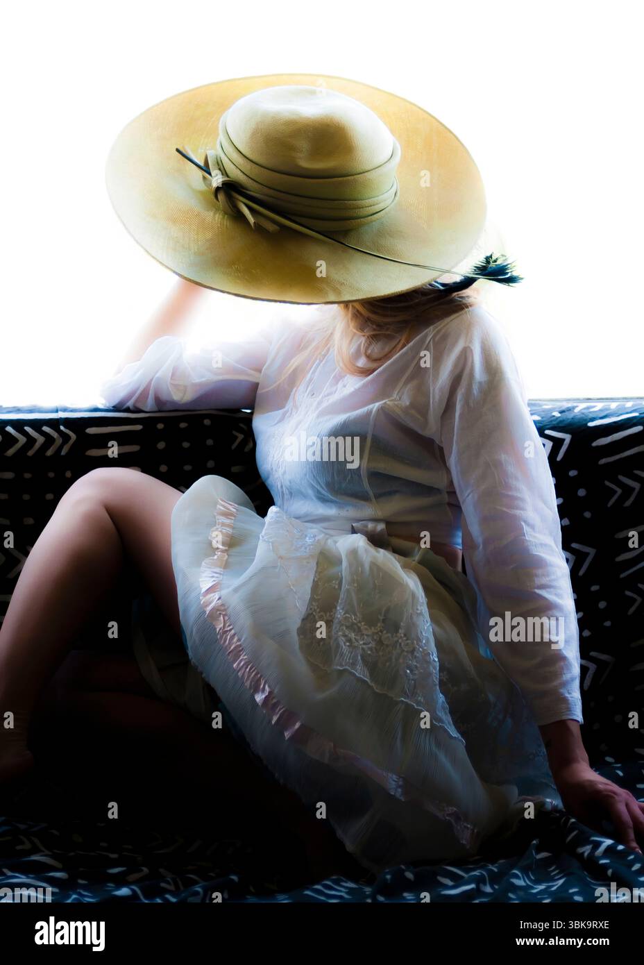 Una donna che indossa un grande cappello e un vestito bianco è seduta su un divano. Foto Stock