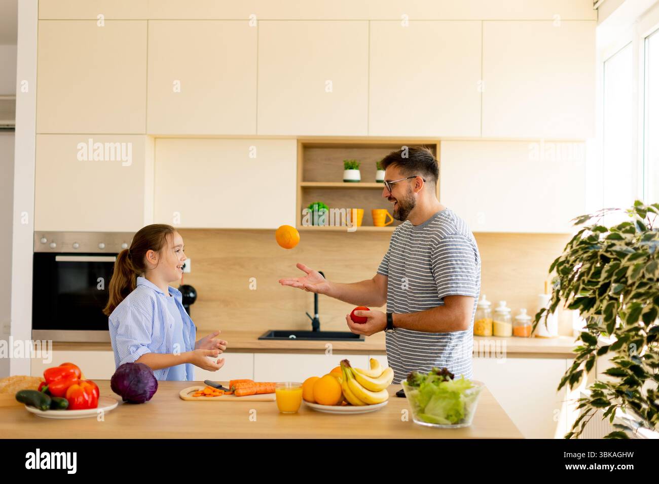Un padre e una figlia condividono risate e divertimento mentre destreggiano frutti vivaci nella loro cucina moderna, evidenziando la gioia di cucinare insieme. Foto Stock