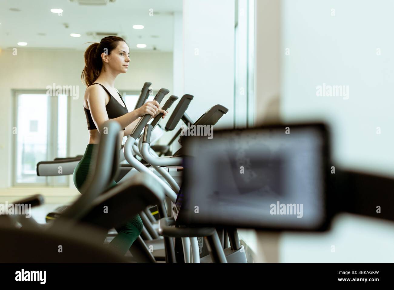 Una persona concentrata si impegna in un esercizio aerobico su un'ellittica, circondata da un'atmosfera luminosa e motivante della palestra, migliorando il percorso di fitness. Foto Stock
