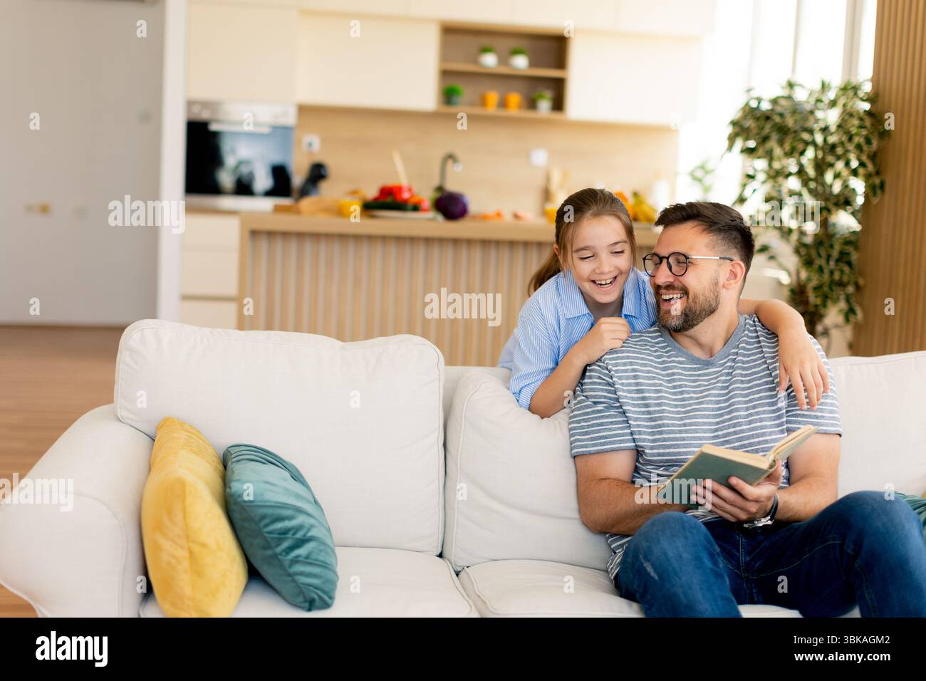 Un padre sorridente condivide una storia deliziosa con sua figlia, seduto su un comodo divano in una casa luminosa e moderna. Foto Stock