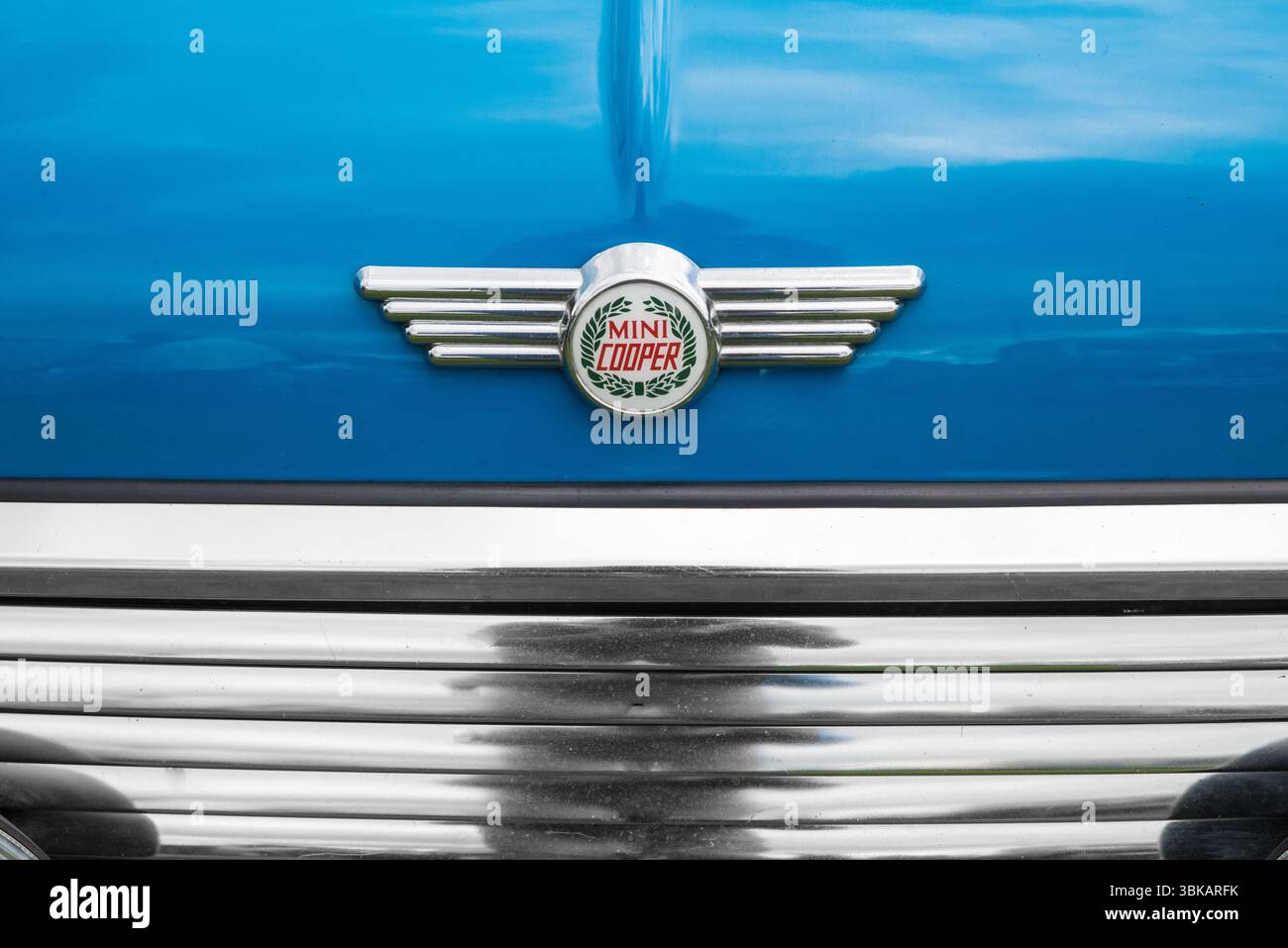 Primo piano di una classica spilla cromata con alette Mini Cooper con il logo della corona d'alloro sul cofano di una Mini Cooper blu Foto Stock