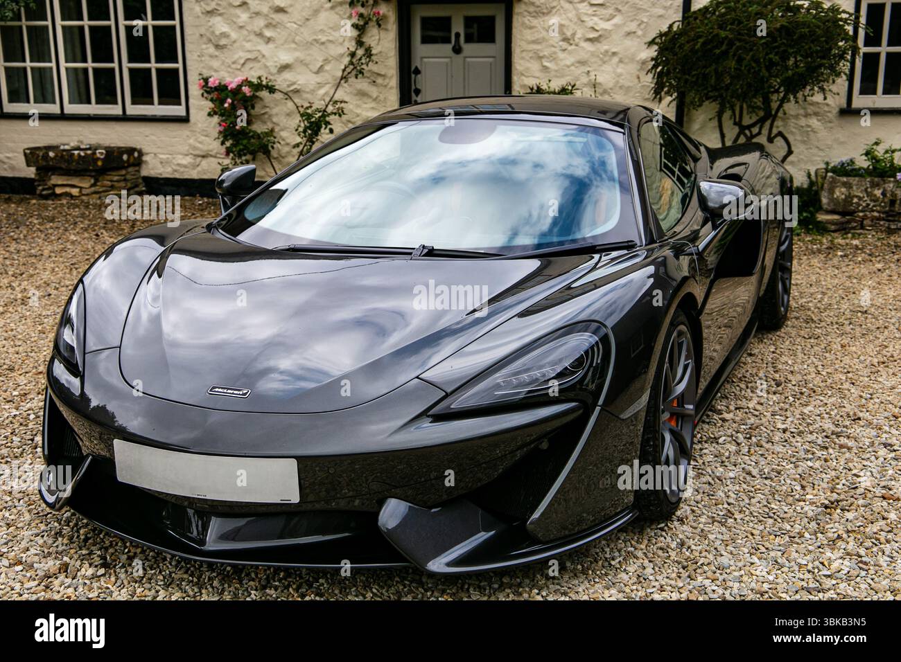 Elegante auto sportiva nera degli anni '570 della McLaren, parcheggiata su un vialetto di ghiaia di fronte a una tradizionale casa di campagna inglese Foto Stock