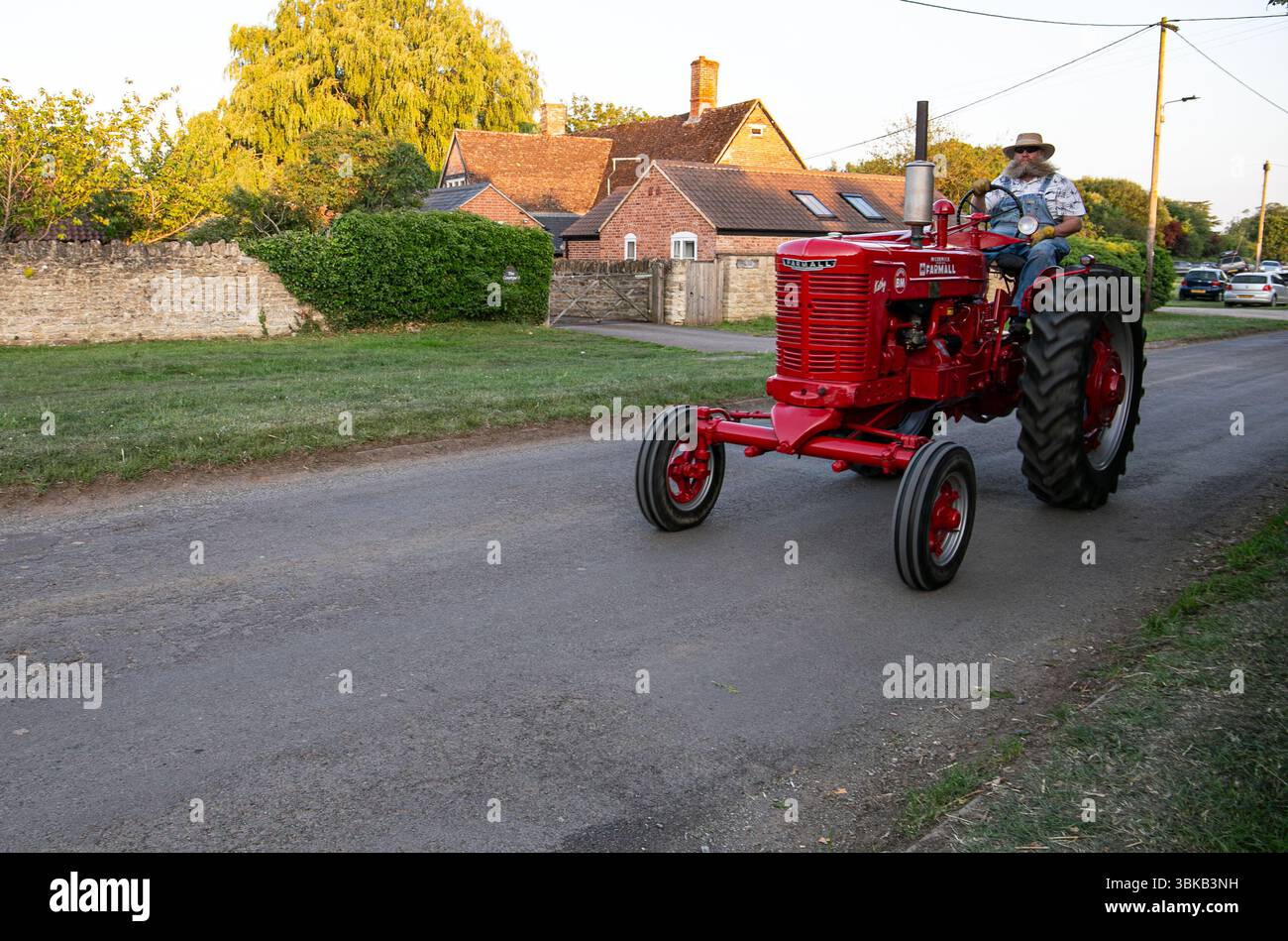 Un uomo che guida un trattore rosso d'epoca al Poulshot Wiltshire veicolo d'epoca che si riunisce con tradizionali case in mattoni sullo sfondo durante il tramonto Foto Stock