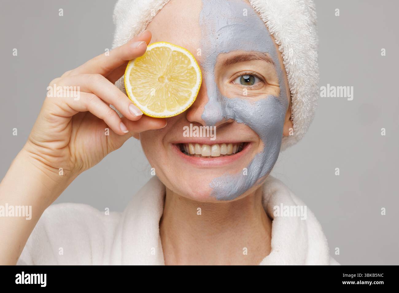 Donna felice che applica la maschera facciale in argilla e tiene in mano il limone, si diverte a rassodare, idratare e anti-invecchiamento a casa. Foto Stock