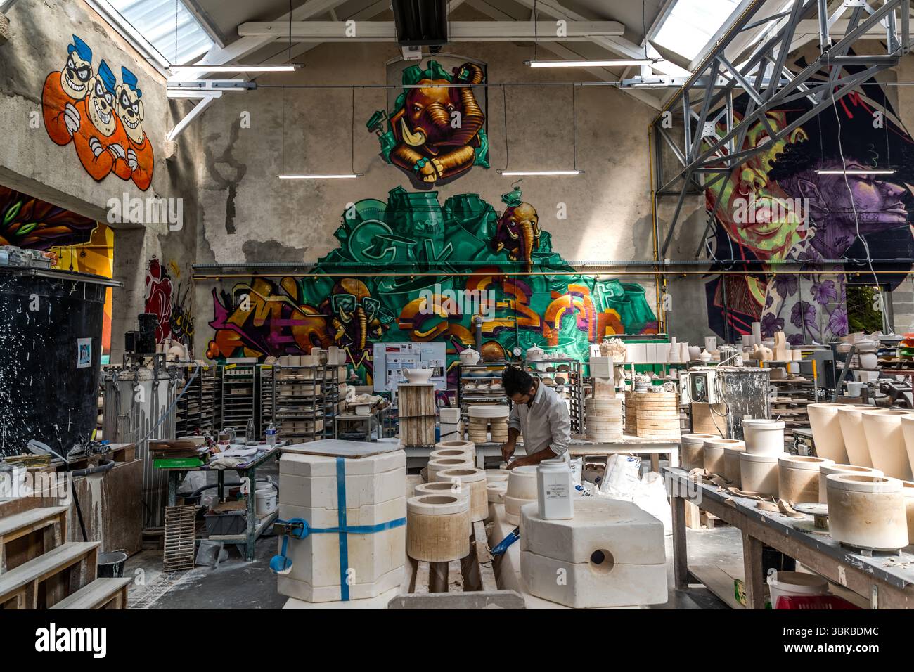 La bottega di porcellana artistica Ateliers Arquié in Avenue Baudin a Limoges, Haute-Vienne, Nouvelle-Aquitaine, Francia, unisce artigianato tradizionale con forme, colori e tecniche moderne. In modo appropriato, un graffiti dell'elefante nel negozio di porcellana adorna la parete. Un artigiano lavora nell'alta sala tra stampi in gesso, scaffali di pezzi grezzi e banchi da lavoro. In alto a sinistra, tre figure con berretti blu sorridono dalla parete, e a destra un grande volto brilla in viola e verde. La bottega è stata fondata nel 1996 e si specializza nella progettazione e produzione di pezzi personalizzabili e artistici Foto Stock
