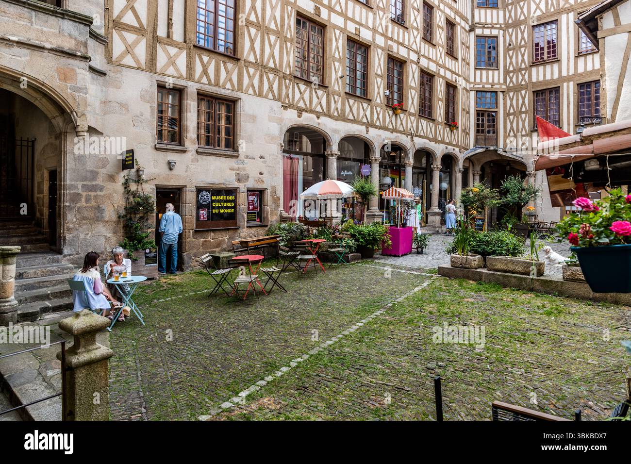 Il cortile della Cour du Temple nella vecchia città di Limoges, Haute-Vienne, Nouvelle-Aquitaine, Francia, ha una storia speciale come luogo di incontro centrale del vecchio quartiere artigianale e commerciale della città. Un corridoio collega il cortile alla Rue du Consulat. Si apre su un imponente insieme di case in granito e in legno a graticcio con arcate e un’elegante scala rinascimentale. Oggi, il cortile ospita piccoli caffè e boutique. Tavoli da caffè e sedie pieghevoli si trovano sul terreno lastricato e ricoperto d’erba tra piante in vaso e ombrelloni. Due donne sono sedute a un tavolo a sinistra. Un uomo è in piedi nel Foto Stock