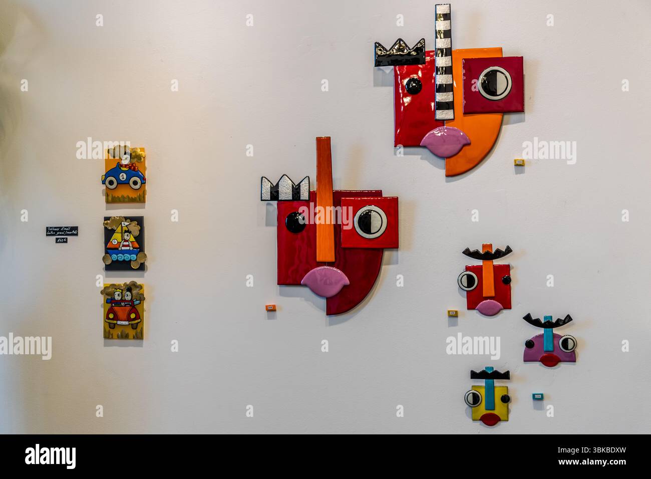 Opere murali moderne smaltate dell'artista Delphine Peytour-Quendolo sono appese a un muro bianco nello studio d'arte e boutique "Le Bocal" sul Boulevard Louis Blanc a Limoges, Haute-Vienne, Nouvelle-Aquitaine, Francia. Due grandi volti rossi con corone bianche e nere, nasi a strisce e bocche rosa hanno un aspetto cubista. Sotto di loro pendono tre piccoli volti in rosso, viola e giallo. A sinistra sono appese tre piccole immagini di un'auto da corsa blu, di una barca a vela e di un'auto rossa, accanto a un piccolo cartello. L'artista lavora con smalto su rame. Le sue figure presentano colori audaci, forme geometriche semplici e occhi sorprendenti. Ena Foto Stock