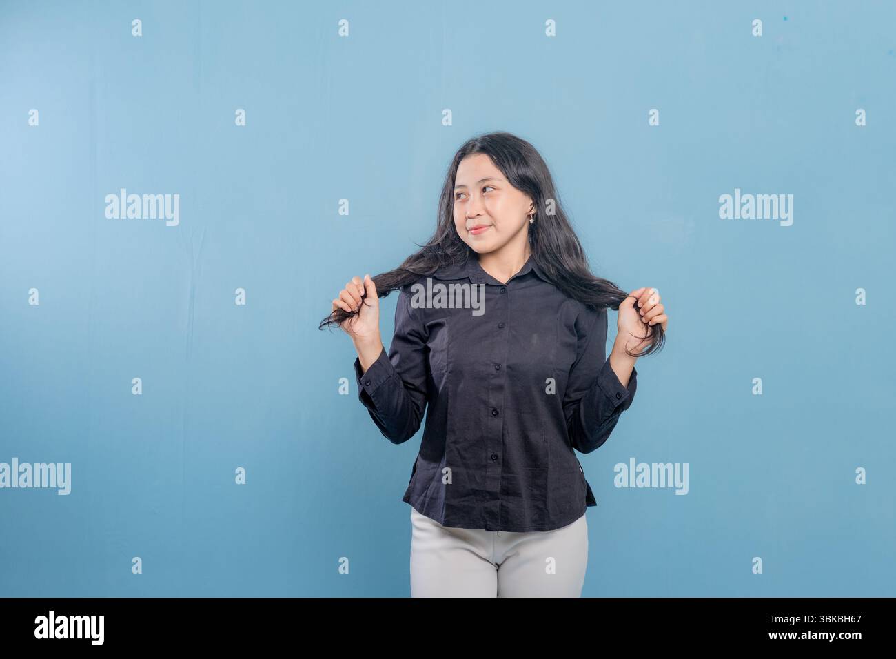 Una donna con i capelli lunghi sorride e si tiene i capelli indietro. Indossa una camicia nera e pantaloni bianchi Foto Stock