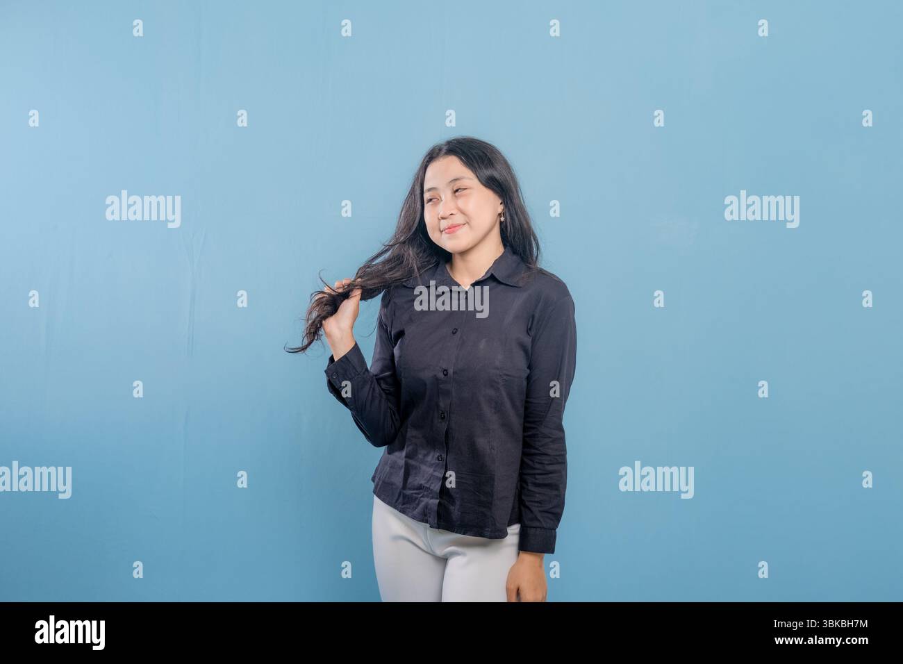 Una donna con i capelli lunghi sorride e si tiene i capelli indietro. Indossa una camicia nera e pantaloni bianchi Foto Stock