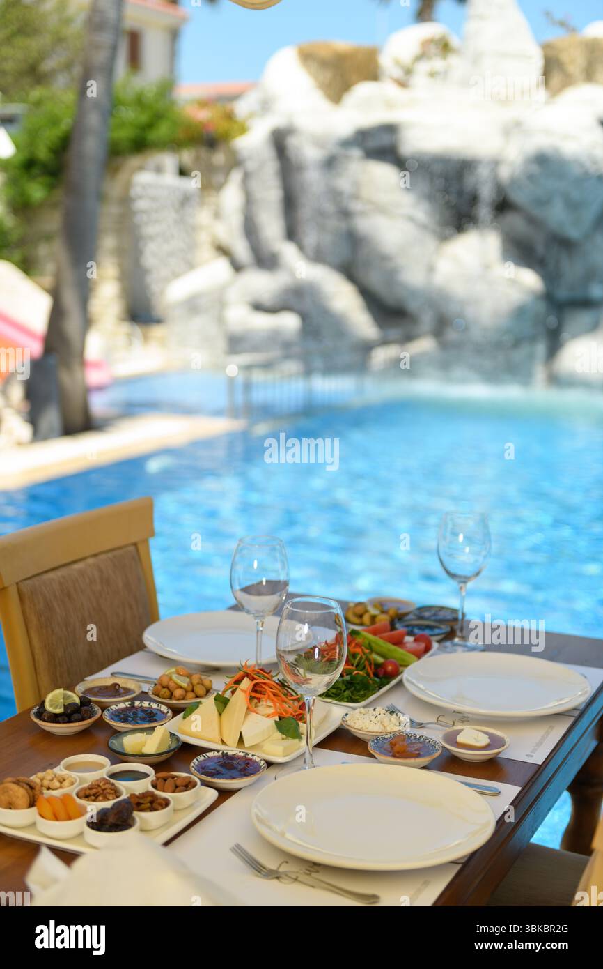 Tavolo in legno per colazione con insalata, formaggio, olive, marmellata, noci, e bicchieri da vino. Piscina sullo sfondo e una singola sedia in legno nella vista. Foto Stock