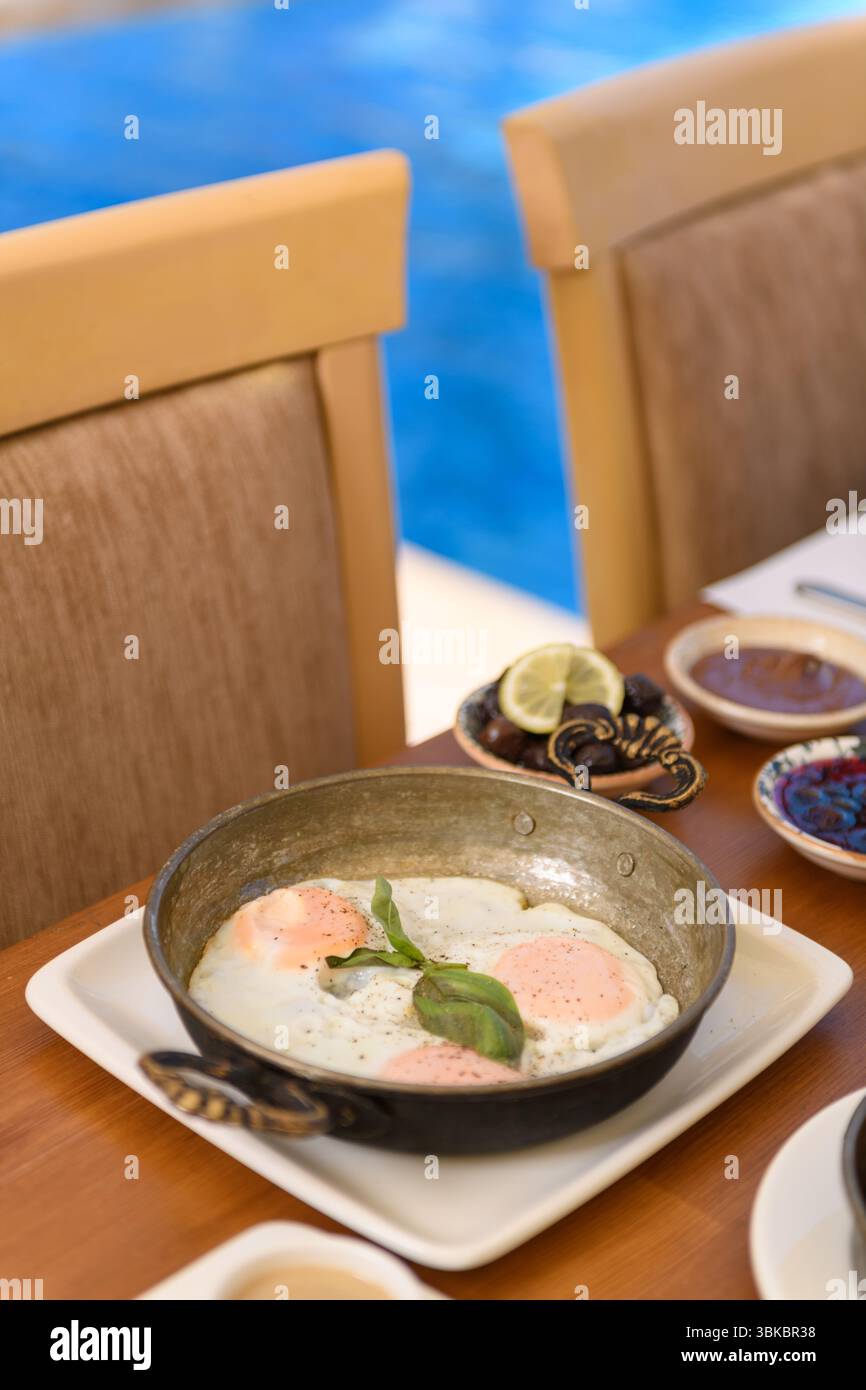 Una tranquilla colazione in hotel con uova fritte su un tavolo di legno, due sedie e una piscina sullo sfondo sotto la luce naturale soffusa Foto Stock