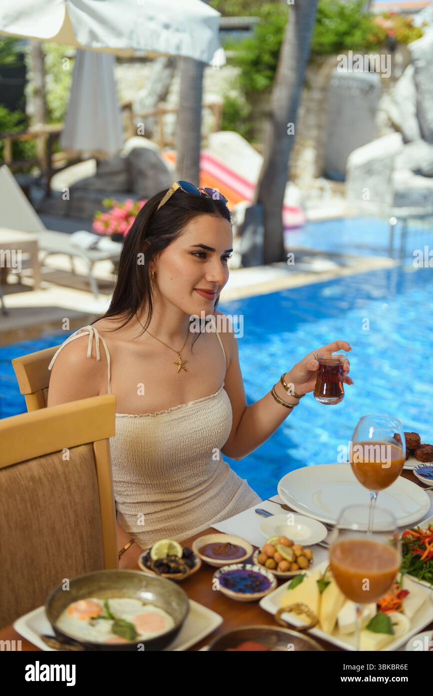 Una donna bruna in un abito beige che tiene in mano un bicchiere di tè durante la colazione in hotel. Formaggio, olive, insalata e succo sul tavolo. Piscina con cascata Foto Stock