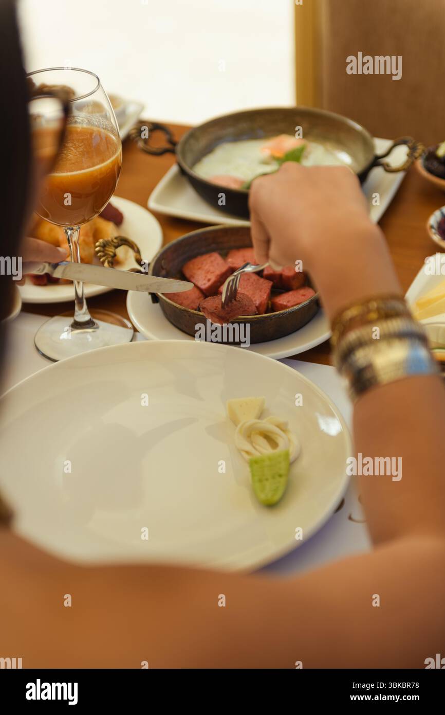 Colazione in hotel con un piatto di uova fritte e salsicce. Una donna usa una forchetta per portare una salsiccia dal piatto di servizio al suo. Accogliente e rilassante Foto Stock