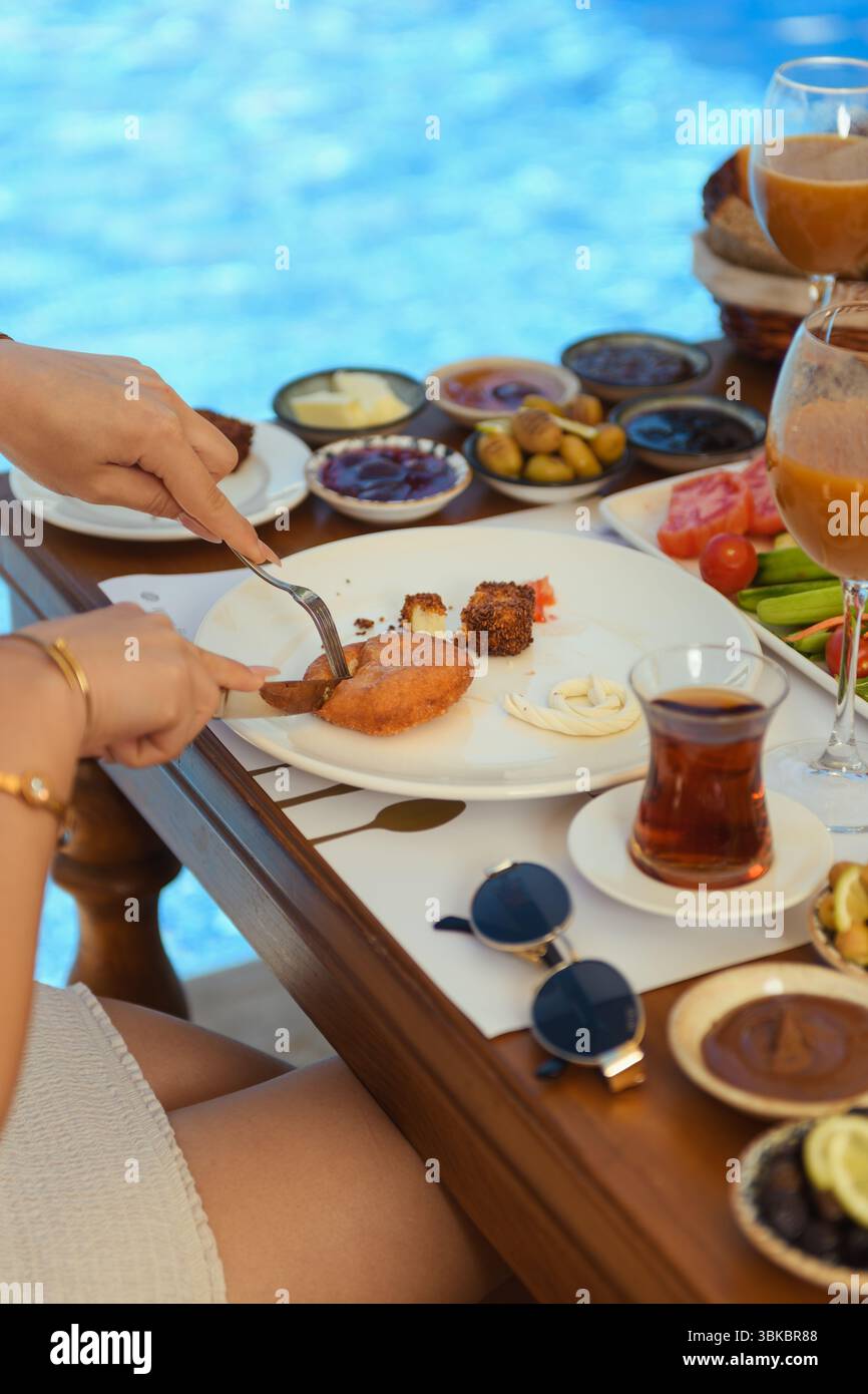 Le mani femminili affettano il cibo su un piatto da colazione al tavolo di un hotel di legno con tè, insalata, olive e occhiali da sole. Atmosfera mattutina rilassata. Foto Stock