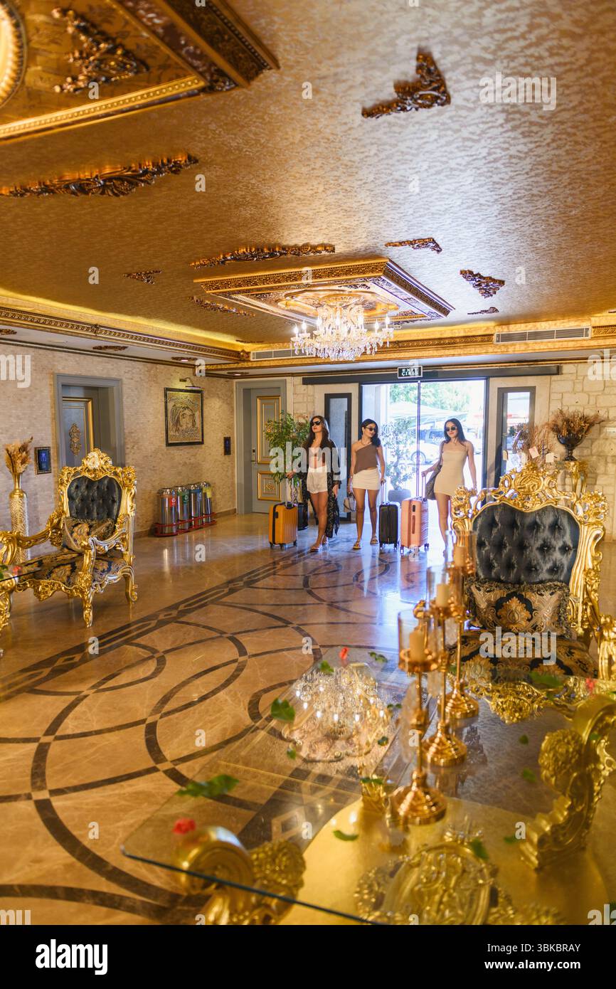 Lobby dell'hotel opulenta in stile barocco con accenti dorati, mobili in velluto blu, superfici in marmo, sontuosi lampadari e tre ospiti femminili in arrivo Foto Stock