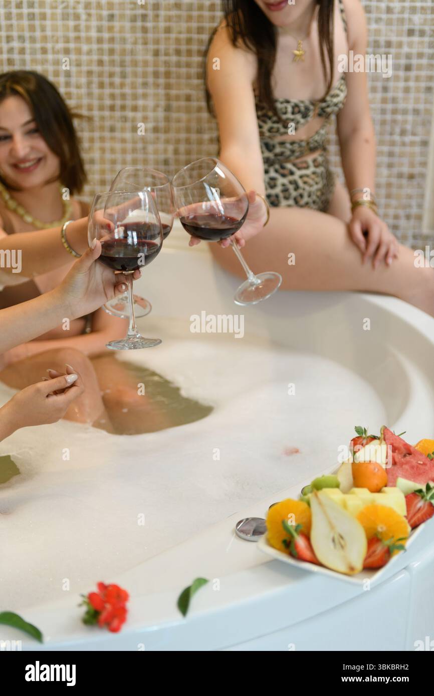 Ragazze allegre che si rilassano in una frizzante jacuzzi, sorseggiano vino e condividono un momento di gioia in un hotel elegante. Foto Stock