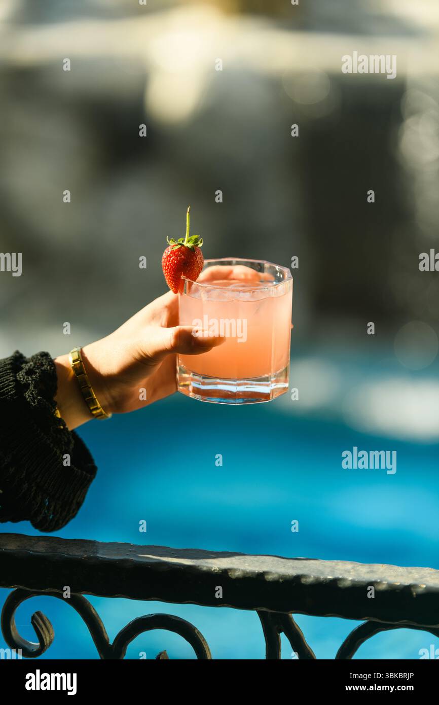 Primo piano della mano di una donna che tiene in mano un vivace cocktail di frutta condito con una fragola, evocando l'estate e rinfrescante. Foto Stock