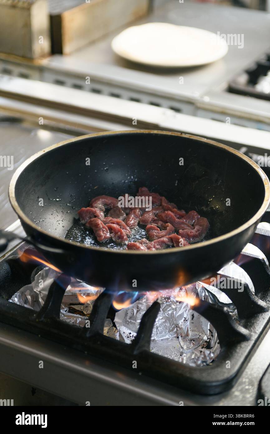 La carne succosa viene cucinata in una padella su una stufa calda nella cucina di un ristorante, catturando il momento della preparazione sfrigolante in un ambiente culinario. Foto Stock