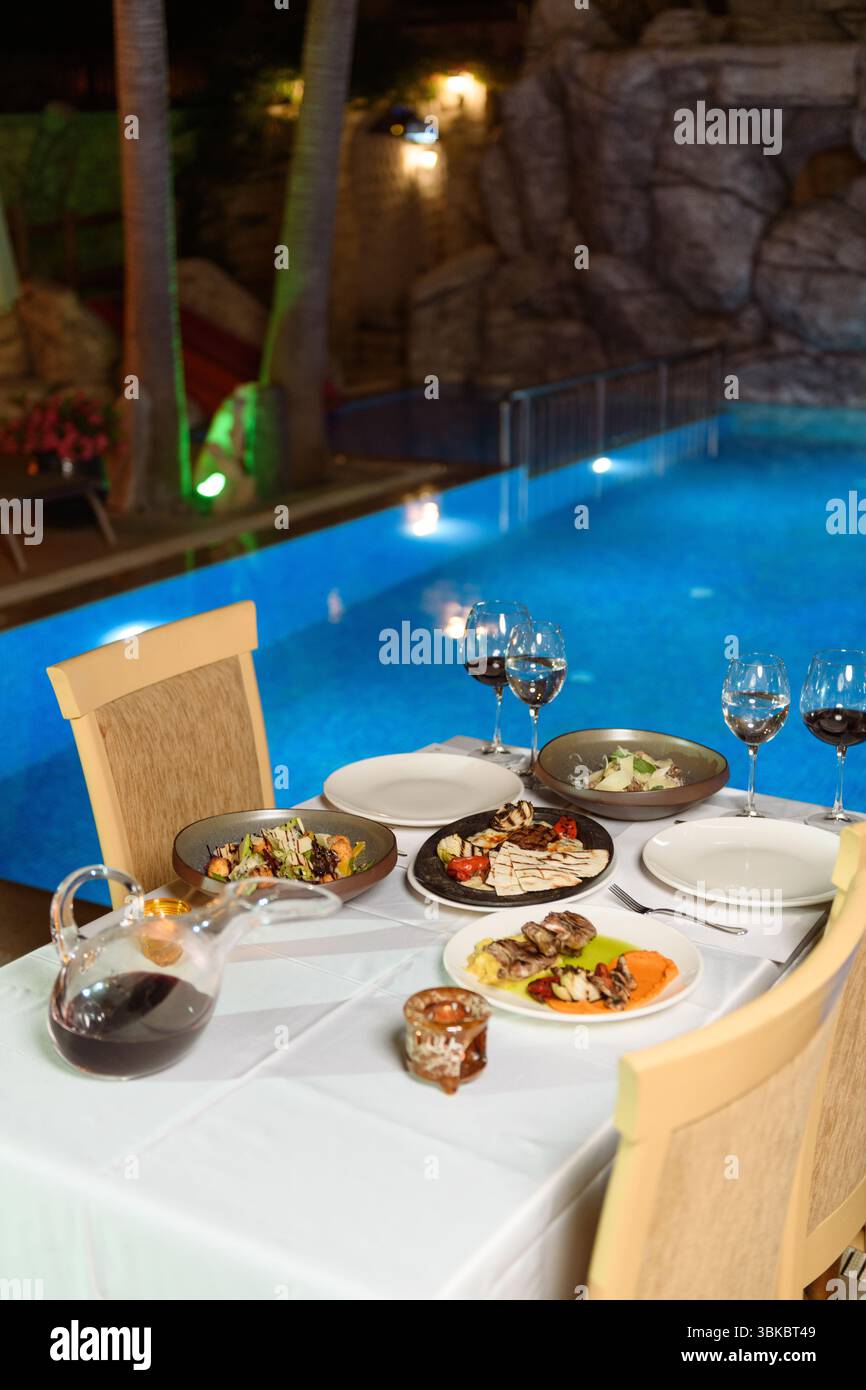 Elegante pasto serale a bordo piscina con bicchieri da vino, carne alla griglia con verdure e illuminazione serale. Foto Stock