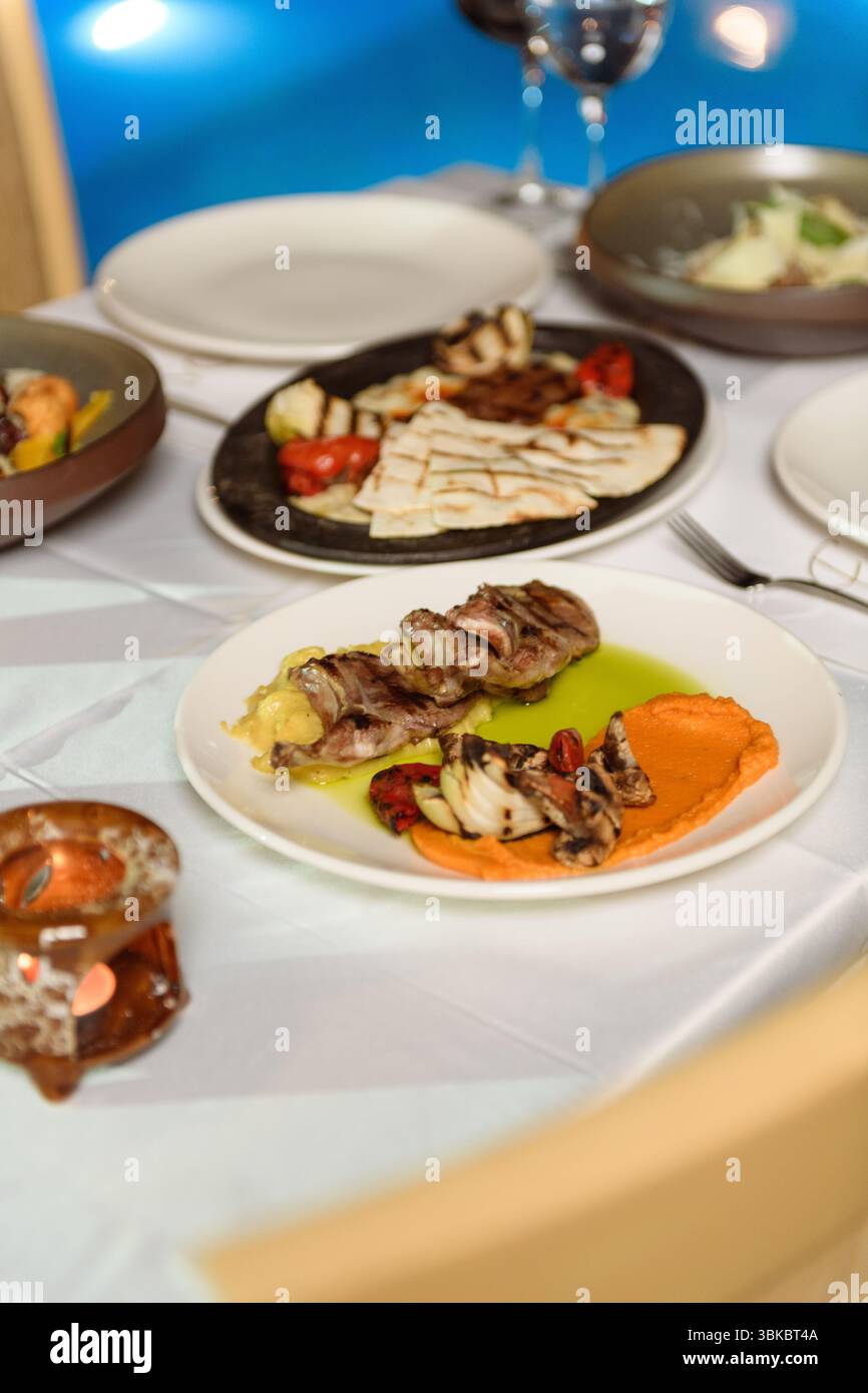 Deliziosi piatti di carne presentati su un tavolo vestito di bianco durante l'elegante cena dell'hotel con vista sulla piscina sullo sfondo. Foto Stock