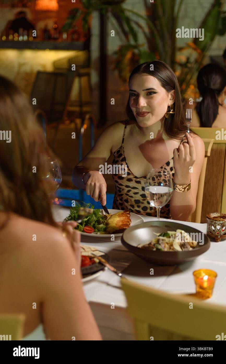 Attraente donna bruna che cena al tavolo del ristorante dell'hotel pieno di vari piatti di carne e insalate, piscina visibile sullo sfondo. Foto Stock