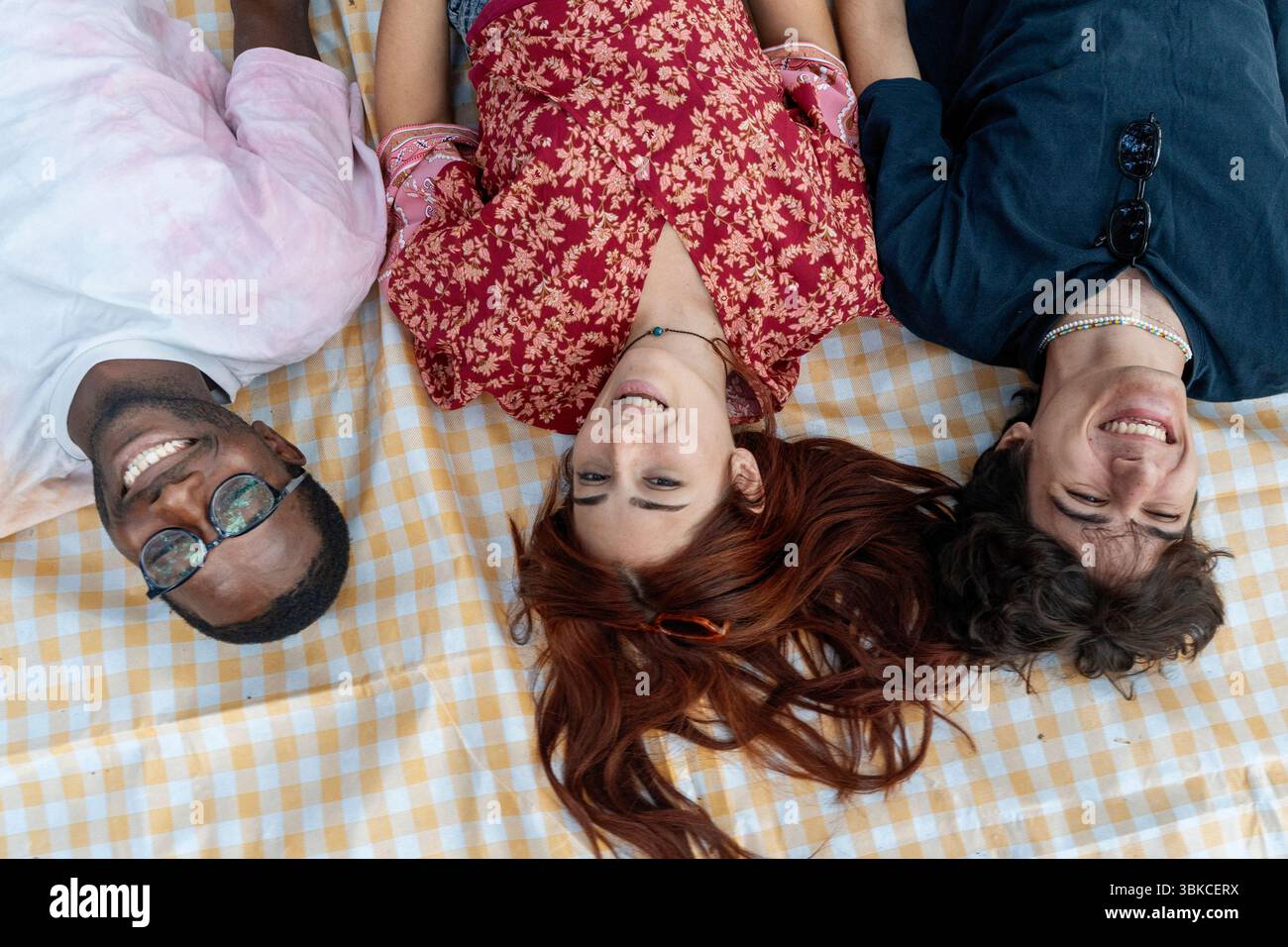 Un gruppo variegato di giovani amici giace con la testa insieme in cerchio su una coperta da picnic, guardando in alto e sorridendo. Concetto di amicizia e unità Foto Stock