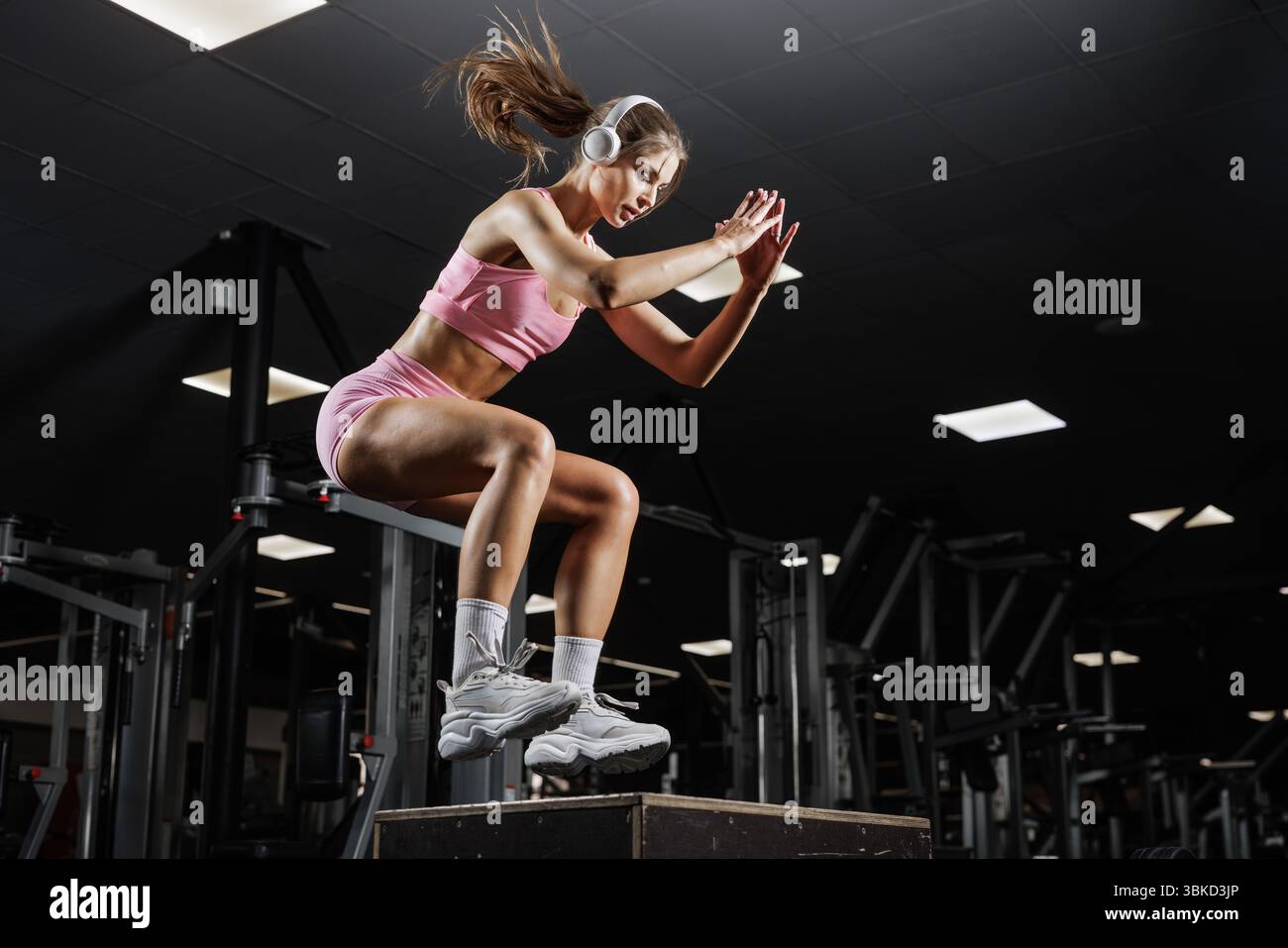 Fit donna che fa box jump in palestra indossando abbigliamento sportivo rosa e cuffie. Catturati a mezz'aria, dimostrando potenza e prestazioni atletiche. Foto Stock