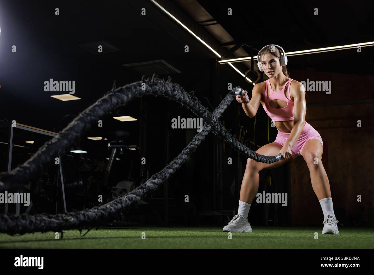Fit Woman in outfit rosa da palestra con corde da battaglia. Potente immagine di allenamento a intervalli ad alta intensità, concentrazione e forza in una palestra buia Foto Stock