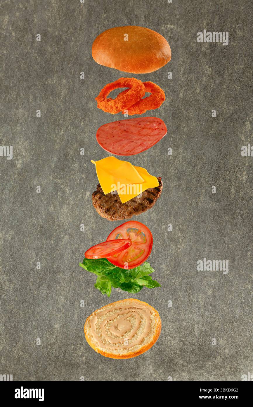 Hamburger volante con i suoi ingredienti: Pane lattuga pomodori formaggio di manzo salame e cipolle croccanti su sfondo grigio. Foto Stock