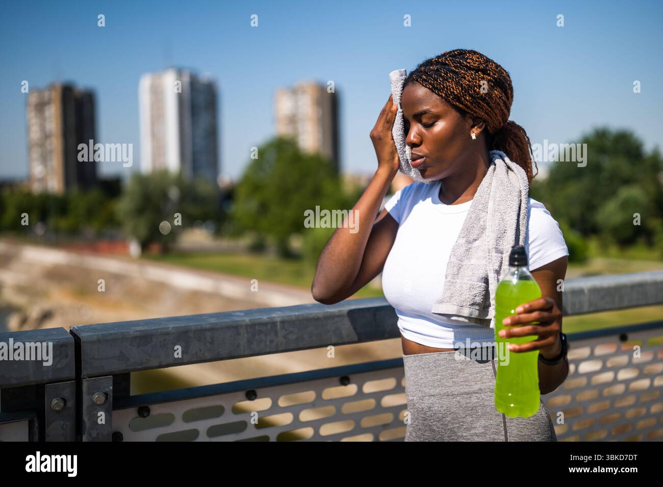 Ritratto di una giovane donna afroamericana in abbigliamento sportivo che pulisce il sudore con un asciugamano e beve una bevanda energetica dopo l'esercizio fisico all'aperto. Foto Stock