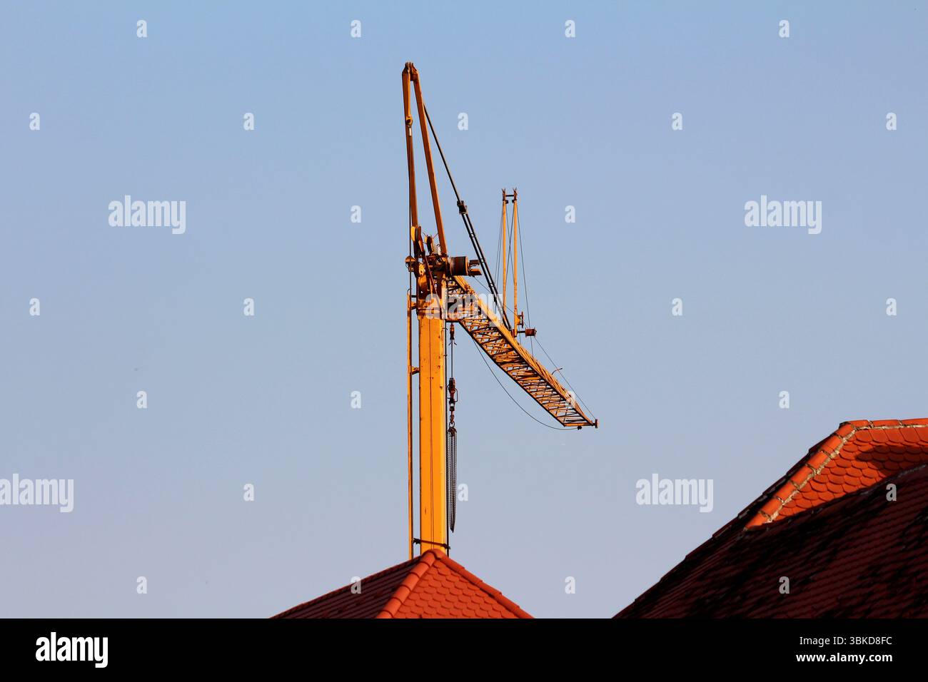 Un'alta gru da costruzione gialla si innalza sopra i tetti di tegole rosse, il suo braccio angolato contro un cielo azzurro limpido, inondato dalla calda luce solare serale durante il periodo Foto Stock