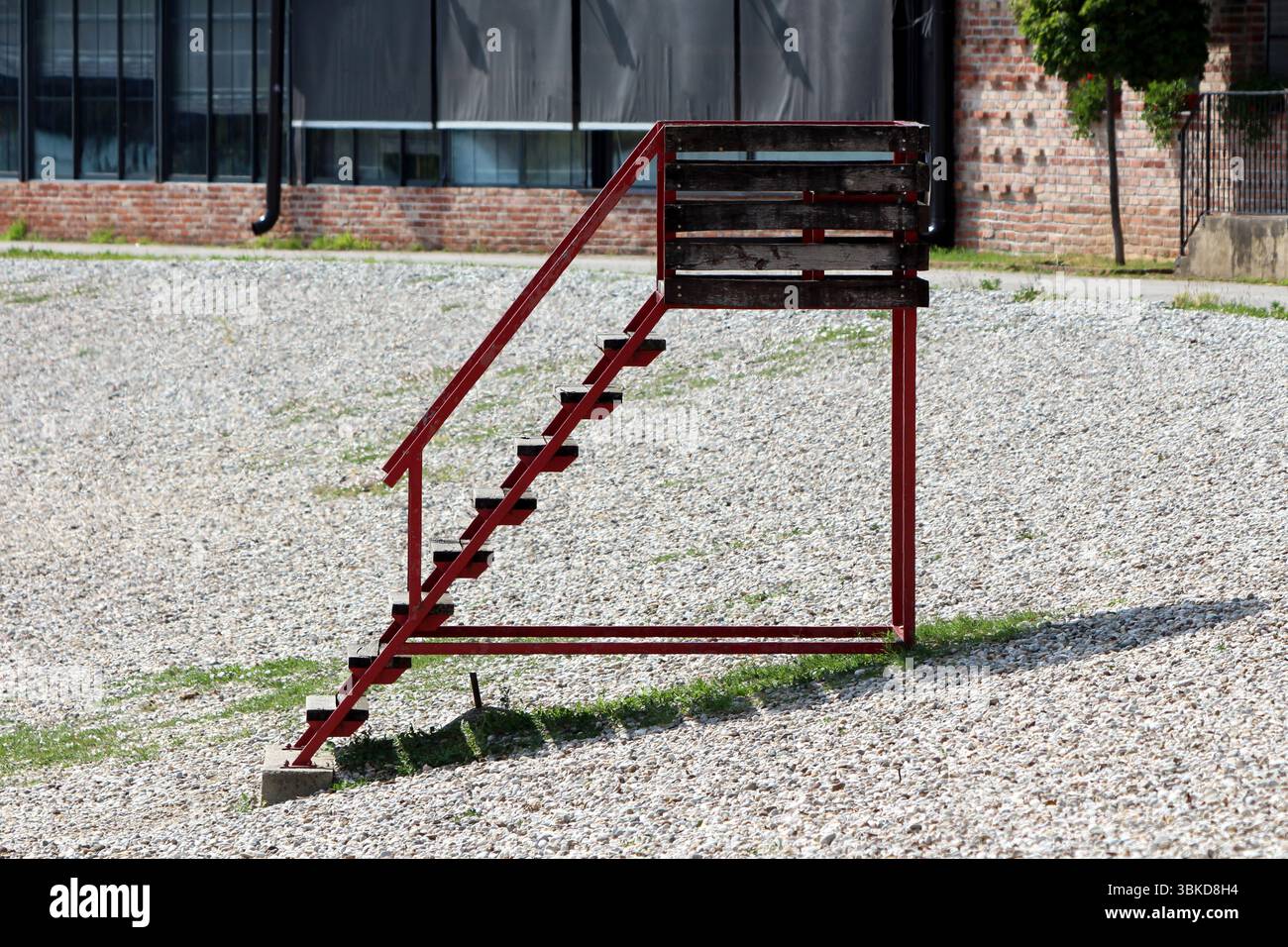 Semplice scala in metallo rosso con una piccola piattaforma in legno si erge da sola su un lotto di ghiaia, gettando un'ombra netta, con mattoni e vetri industriali Foto Stock