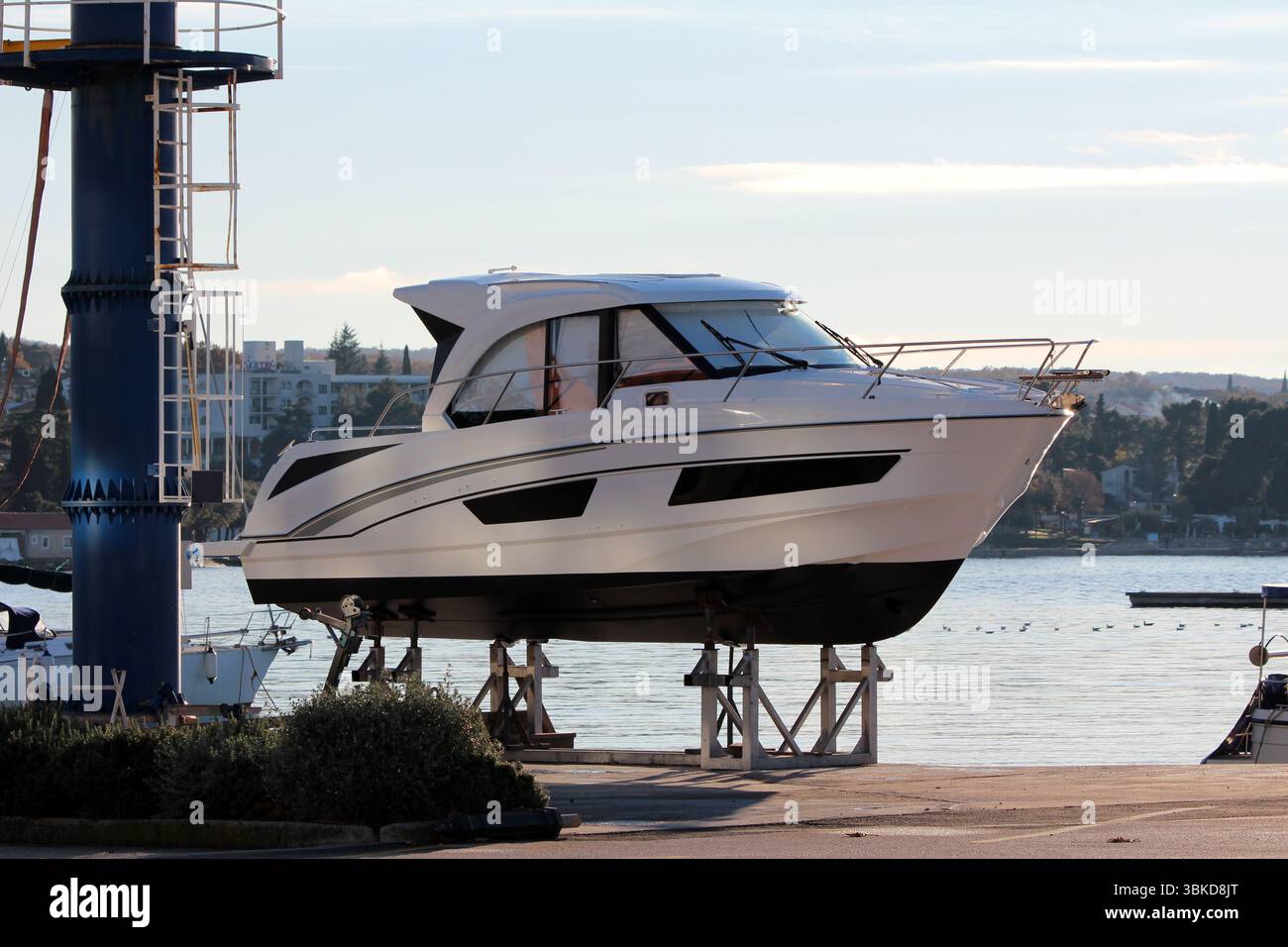Elegante yacht a motore bianco con finestre colorate e accenti neri si erge su supporti metallici per la manutenzione in un porticciolo, con acqua calma e distensione Foto Stock
