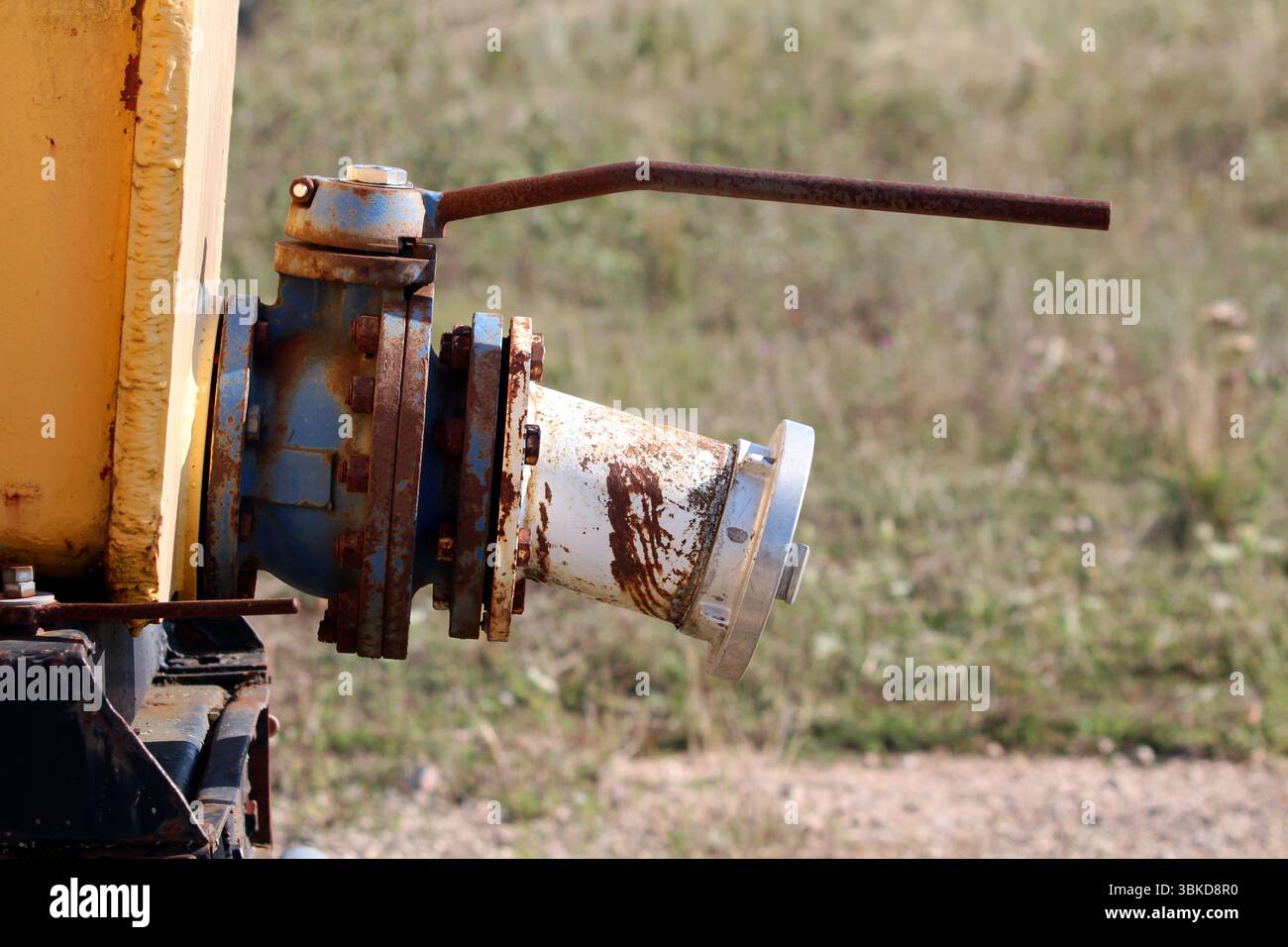 Valvola industriale di grandi dimensioni con raccordi blu e bianchi e una lunga leva arrugginita è fissata ai macchinari gialli, mostrando corrosione e usura, impostata all'esterno Foto Stock